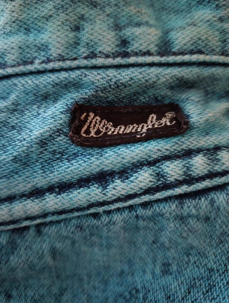 May include: Close-up of a blue denim fabric with a black leather patch that says "Wrangler" in white lettering.