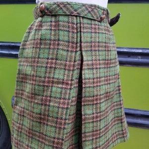Peut inclure: Une jupe en tissu écossais vert et marron avec un devant plissé et un bouton marron à la taille.