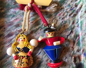 May include: Two wooden figurines, a girl and a boy, are hanging from a red and white cord. The girl is wearing a yellow dress with red and white accents and has yellow hair. The boy is wearing a blue and red outfit and has a black hat. The figurines are hanging from a wooden bar.