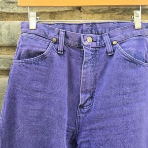 Może przedstawiać: Zbliżenie pary fioletowych dżins&oacute;w. Dżinsy wiszą na drewnianym wieszaku. Dżinsy mają zapięcie na guziki i kieszenie. Denim ma teksturowany wygląd.