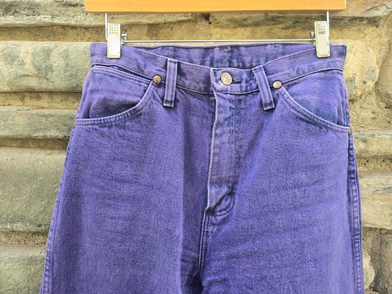 Może przedstawiać: Zbliżenie pary fioletowych dżins&oacute;w. Dżinsy wiszą na drewnianym wieszaku. Dżinsy mają zapięcie na guziki i kieszenie. Denim ma teksturowany wygląd.