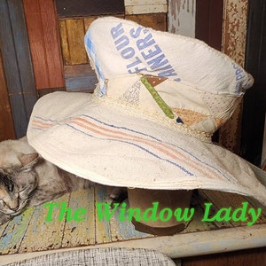 May include: A large, white sun hat made from repurposed fabric with blue and pink stripes. The hat is sitting on a wooden table with a gray and white cat sleeping underneath it. The text "The Window Lady" is visible on the table.