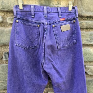 Może przedstawiać: Zbliżenie na tył fioletowych dżins&oacute;w Wrangler. Dżinsy mają dwie tylne kieszenie z wszytym logo Wrangler. Widoczna jest mała czerwona metka z logo Wrangler. Dżinsy wiszą na metalowym wieszaku.