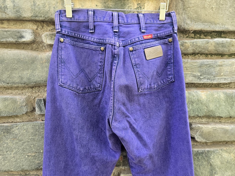 Może przedstawiać: Zbliżenie na tył fioletowych dżins&oacute;w Wrangler. Dżinsy mają dwie tylne kieszenie z wszytym logo Wrangler. Widoczna jest mała czerwona metka z logo Wrangler. Dżinsy wiszą na metalowym wieszaku.
