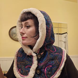 Puede incluir: Un calentador de cuello con capucha de color azul y rosa con estampado paisley, ribeteado con piel sintética blanca. El calentador de cuello tiene dos botones negros.