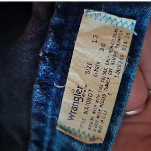 May include: Wrangler brand jeans with a white tag that says "STYLE NA7080T SIZE LENGTH 13 36 MACHINE WASH WITH LIKE COLORS, ONLY NON-CHLORINE BLEACH WHEN NEEDED. TUMBLE DRY. REMOVE PROMPTLY. C 1JAEV349 814 26 MADE IN U.S.A."
