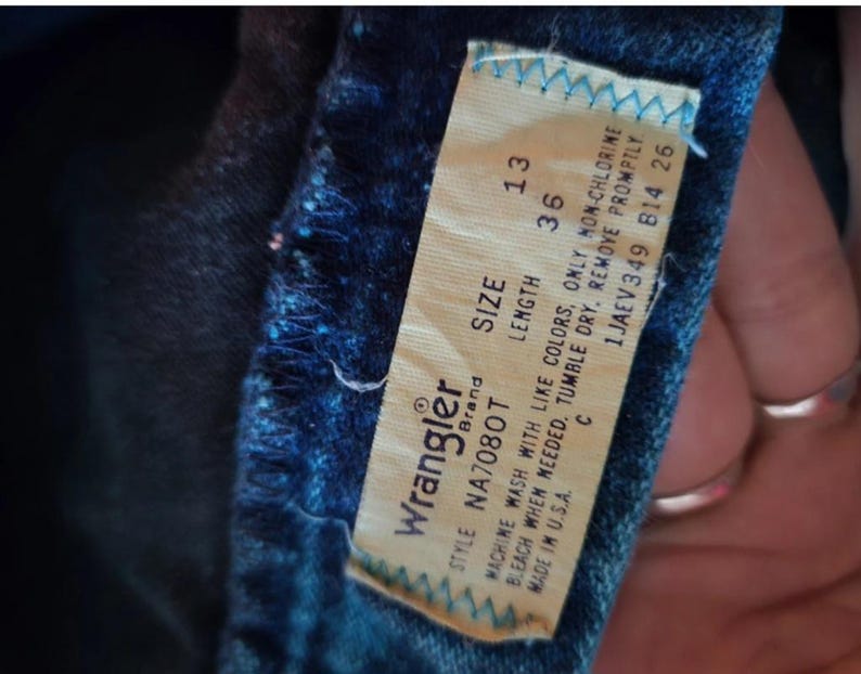 May include: Wrangler brand jeans with a white tag that says "STYLE NA7080T SIZE LENGTH 13 36 MACHINE WASH WITH LIKE COLORS, ONLY NON-CHLORINE BLEACH WHEN NEEDED. TUMBLE DRY. REMOVE PROMPTLY. C 1JAEV349 814 26 MADE IN U.S.A."