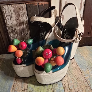 May include: Platform sandals with a white woven base, teal blue accents, and beige straps. The sandals are adorned with colorful, artificial fruit. The heels have a beige woven texture. The sandals have the text "for Kim" on the insole.