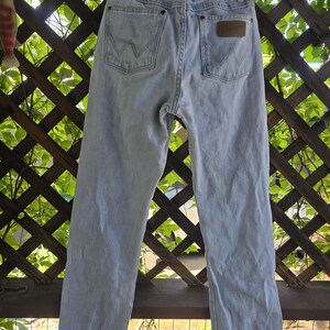 May include: Light blue denim jeans featuring a high-waisted design with belt loops, a metal button closure, and two back pockets. Each back pocket displays a signature W-shaped stitching pattern in light thread, with a brown leather patch.