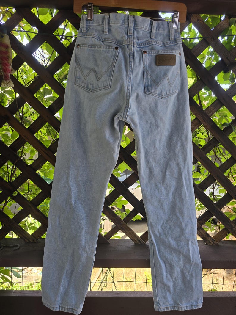 May include: Light blue denim jeans featuring a high-waisted design with belt loops, a metal button closure, and two back pockets. Each back pocket displays a signature W-shaped stitching pattern in light thread, with a brown leather patch.