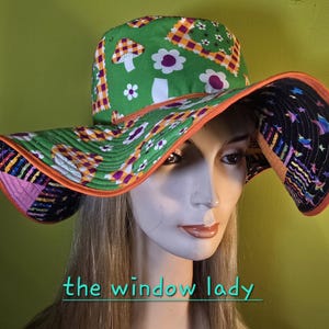 May include: A wide-brimmed sun hat with a green, orange, and purple floral and geometric pattern. The hat features a mushroom and flower design on the crown and brim. The brim is lined with a contrasting black fabric with colorful star shapes. The text "the window lady" is visible.