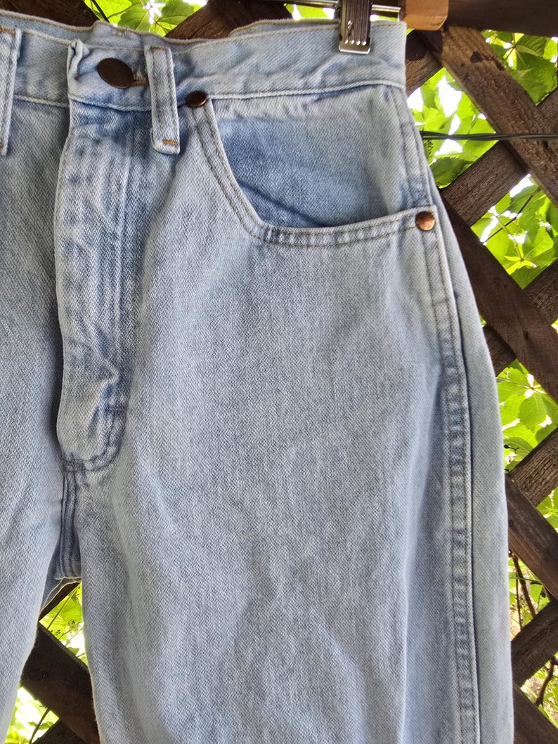 May include: Light blue denim jeans featuring a high-waisted design with a button fly closure, copper-toned metal rivets at the pocket corners, and a classic five-pocket construction. The fabric has a light wash finish with visible stitching.