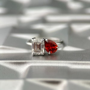 May include: A silver ring with a square-shaped clear gemstone and a teardrop-shaped red gemstone.