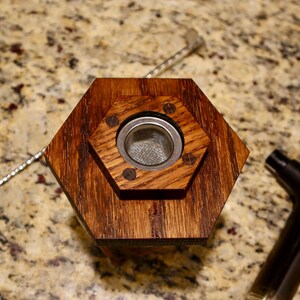 Pu&ograve; includere: Una base esagonale in legno con un inserto in rete metallica. La base &egrave; tinta di un colore marrone scuro e ha una finitura liscia. L'inserto in rete metallica &egrave; circolare e ha una maglia fine.