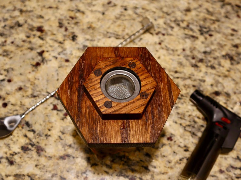 Pu&ograve; includere: Una base esagonale in legno con un inserto in rete metallica. La base &egrave; tinta di un colore marrone scuro e ha una finitura liscia. L'inserto in rete metallica &egrave; circolare e ha una maglia fine.