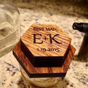 Pu&ograve; includere: Un sottobicchiere esagonale in legno con inciso il testo "Best Man E+K 1.19.2015". Il sottobicchiere &egrave; posto sopra un bicchiere con un liquido marrone.