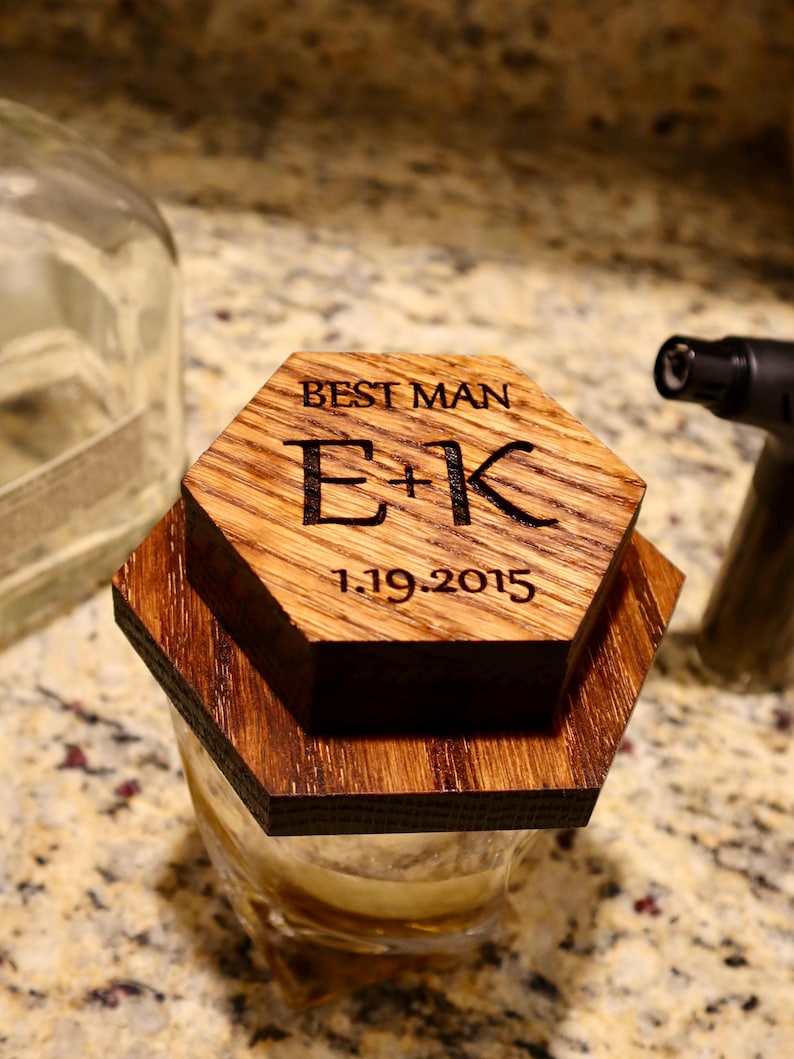 Pu&ograve; includere: Un sottobicchiere esagonale in legno con inciso il testo "Best Man E+K 1.19.2015". Il sottobicchiere &egrave; posto sopra un bicchiere con un liquido marrone.