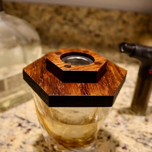Pu&ograve; includere: Un coperchio in legno esagonale con un'apertura rotonda al centro &egrave; posto sopra un bicchiere di vetro trasparente riempito di un liquido marrone chiaro. Il coperchio &egrave; in legno scuro con una venatura naturale.