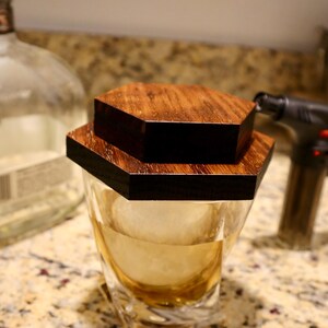 Pu&ograve; includere: Un bicchiere di whisky in vetro trasparente con un coperchio in legno esagonale. Il bicchiere &egrave; pieno di liquido ambrato e ghiaccio. Il coperchio &egrave; composto da due pezzi di legno, uno sopra l'altro.