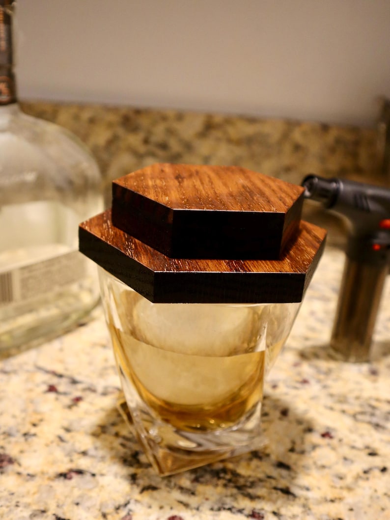 Pu&ograve; includere: Un bicchiere di whisky in vetro trasparente con un coperchio in legno esagonale. Il bicchiere &egrave; pieno di liquido ambrato e ghiaccio. Il coperchio &egrave; composto da due pezzi di legno, uno sopra l'altro.