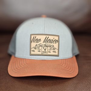 May include: A baseball cap with a light blue crown, a brown brim, and a brown patch. The patch features the words "New Mexico Las Cruces 1912" in a decorative font, with a graphic of a bird and a banner.