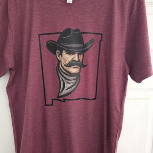 May include: Burgundy t-shirt with a graphic of a cowboy with a mustache, hat, and scarf, set within a black state outline. The design is on the front of the short-sleeved crew neck shirt.