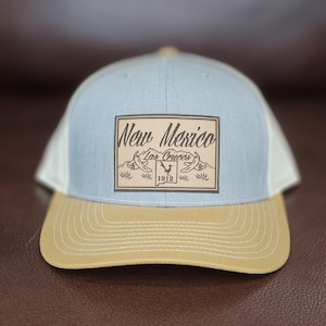 Può includere: Cappellino da baseball azzurro e beige con retro in rete bianca. La parte anteriore presenta una toppa rettangolare marrone con le parole "New Mexico Las Cruces 1912" in stile vintage.