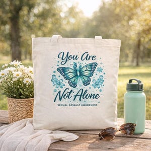 Pu&ograve; includere: Borsa tote color crema con la scritta "You Are Not Alone" e un disegno di farfalla turchese. La borsa &egrave; su un tavolo di legno con una bottiglia d'acqua, occhiali da sole e un cesto di fiori. &Egrave; visibile anche la frase "Sexual Assault Awareness".