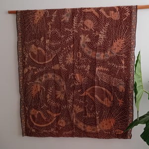 Op de afbeelding: Een bruin en beige batiktextiel met een ingewikkeld patroon van wervelende lijnen en gestileerde wezens. De stof is opgehangen aan een houten staaf, waarbij het gedetailleerde ontwerp en de kleurvariaties worden benadrukt.