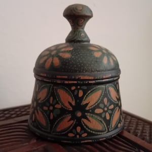 Könnte beinhalten: Ein kleiner, runder Holzbehälter mit Deckel, der eine schwarze Grundfarbe mit handgemalten Blumenmustern in gedämpftem Orange und Salbeigrün aufweist. Der Deckel hat einen passenden Knaufgriff und eine punktierte Textur.