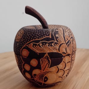 May include: A carved wooden apple-shaped container featuring a dark brown finish with intricate light tan patterns. The surface displays detailed etchings of fish, circular motifs, and floral elements, topped with a curved wooden stem.