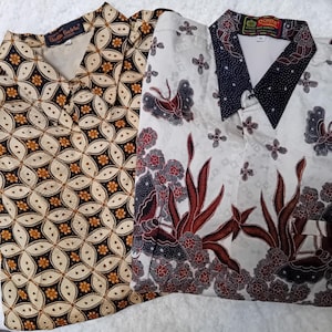 Op de afbeelding: Twee overhemden met lange mouwen met een batikpatroon. Een overhemd heeft een witte achtergrond met bruine en rode bloemenmotieven en vlinders. Het andere overhemd heeft een zwarte achtergrond met een geometrisch patroon van witte en bruine cirkels.