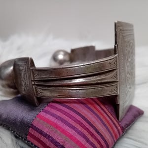 May include: A silver-toned cuff bracelet with intricate etched designs. The bracelet features a rectangular clasp and a rounded end. It rests on a small, colorful pillow with pink and purple stripes, set against a white, fluffy background.
