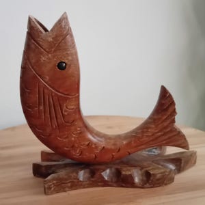 Op de afbeelding: Een bruin gesneden houten vissculptuur met een open mond en een zwart oog, gemonteerd op een horizontale houten basis. Het vissenlichaam heeft gegraveerde schubpatronen en vin-details, terwijl de basis verschillende kleine ronde inkepingen bevat.