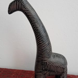 May include: A tall, decorative giraffe sculpture with a black and white chevron pattern. The giraffe has a long neck and stands on four legs. The sculpture is made of a dark material and is displayed on a red patterned surface.
