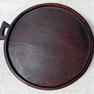 Op de afbeelding: Een ronde, donkerbruine houten dienblad met een opstaande rand en een handvat. De lade heeft een glad oppervlak met zichtbare houtnerf en een donkere vlek. De handgreep is vierkant met een uitgesneden ontwerp. De lade wordt waarschijnlijk gebruikt om te serveren of uit te stallen.