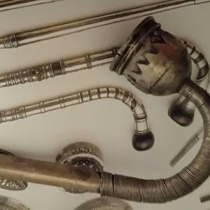 May include: Close-up of antique metal smoking pipes. The pipes have intricate designs and curved stems. The main pipe has a large bowl with a zigzag pattern. The pipes are silver and dark gray.