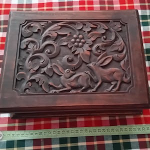 Può includere: Scatola rettangolare in legno con coperchio intagliato con lepri e motivi floreali. La scatola è tinta con un colore marrone caldo. L'intaglio intricato è posto su uno sfondo a scacchi. La scatola è lunga circa 30 cm.