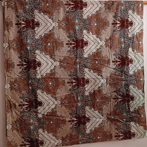Puede incluir: Un textil rectangular con un patrón repetitivo de formas geométricas blancas, marrones y burdeos. La tela está drapeada sobre una barra de madera, mostrando el intrincado diseño y las variaciones de color. La estética general es terrosa y detallada.
