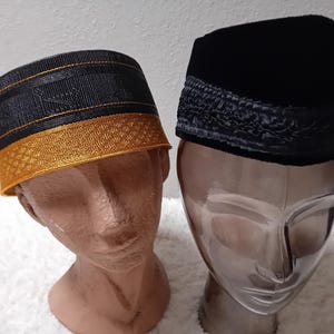 Puede incluir: Dos sombreros kufi tradicionales. Uno es negro con ribete dorado, el otro es negro con una banda decorativa. Ambos se exhiben sobre cabezas de maniquíes sobre un fondo blanco. Los sombreros son probablemente para uso religioso o cultural.