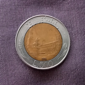 Puede incluir: Una moneda italiana de 500 liras en plata y oro. La moneda presenta un relieve detallado de un edificio y una estatua. El borde de la moneda tiene un diseño similar al trigo. La moneda está fechada en 1989.