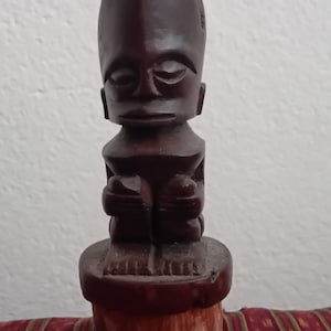 Könnte beinhalten: Eine dunkelbraune Holzstatue einer sitzenden Figur mit großem Kopf und stilisierten Merkmalen. Die Figur sitzt auf einem runden Sockel und die Statue hat eine glatte, polierte Oberfläche. Die Statue ist wahrscheinlich ein Dekorationsgegenstand.