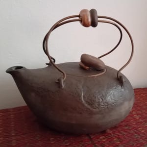 Puede incluir: Una tetera de metal martillado marrón oscuro con un pico curvo y una tapa con bisagras. El asa es de metal y madera, con dos cuentas de madera en la parte superior. La tetera está sobre una superficie estampada roja.