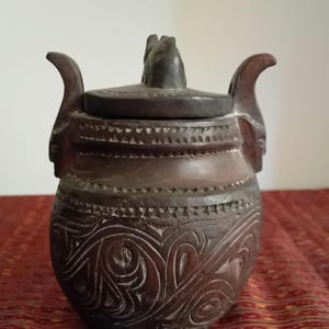 Peut inclure: Un pot en bois sculpté à la main, de couleur marron foncé, avec un couvercle. Le pot présente des motifs tourbillonnants complexes et des poignées décoratives. Le couvercle a une petite figurine sculptée sur le dessus. Le pot est probablement utilisé pour le stockage.