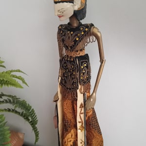 Op de afbeelding: Een gedetailleerde houten pop met een geschilderd gezicht, versierd met een hoge, sierlijke hoofdtooi in tinten bruin, oranje en goud. De pop draagt een zwart-bruin patroon outfit met gouden accenten en een patroon rok. De pop staat rechtop.