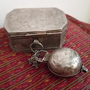 May include: An octagonal silver-toned metal box featuring intricate floral engravings on the lid and sides. A small, hammered silver-toned spherical container is attached to the front of the box by a short metal chain and a circular ring.