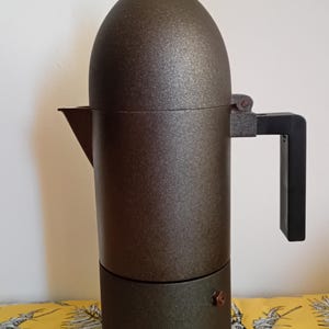 May include: A bronze-colored stovetop espresso maker with a black handle and a black knob on top. The coffee maker has a cylindrical body and a spout for pouring. The espresso maker is a kitchen appliance.