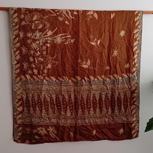 Pode incluir: Um tecido castanho com um padrão detalhado de pássaros, flores e formas geométricas. O tecido é drapeado sobre uma vara de madeira, mostrando o seu design intrincado e variações de cor. As bordas do tecido são bordadas com uma guarnição decorativa.