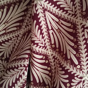 Pode incluir: Close-up de um tecido com um padrão repetitivo de folhas e elementos florais brancos estilizados sobre um fundo bordô escuro. Pequenas lantejoulas douradas estão espalhadas por todo o design.
