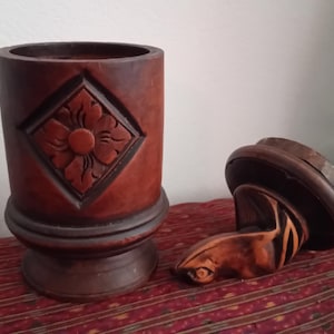 Peut inclure: Deux objets en bois sculpté brun foncé. L'un est un récipient cylindrique avec un motif floral en forme de losange. L'autre est un objet décoratif et incurvé avec une base circulaire. Les objets sont sur une surface à motifs.